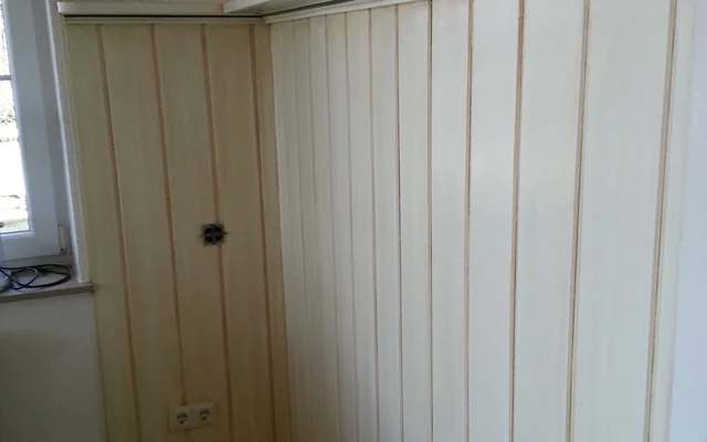 Weiße Holzvertäfelung an zwei Wänden einer Zimmerecke mit einem kleinen Fenster und einer Steckdose an der linken Wand.