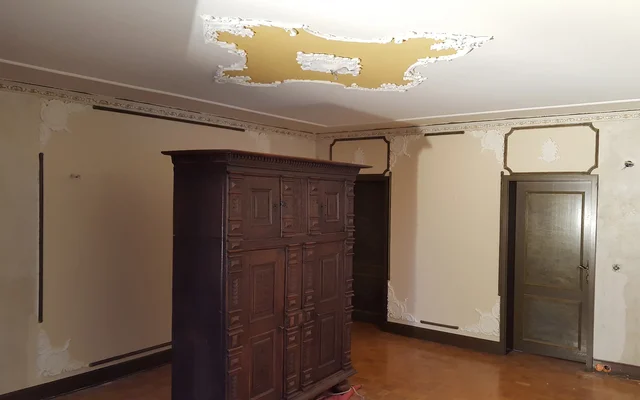 Verlassener Raum mit teilweise beschädigten Wänden und Decke, darin ein großer, dunkler Holzschrank und zwei geschlossene Türen.