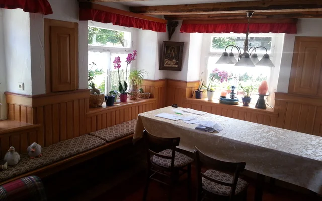 Gemütlicher Essbereich mit Holzvertäfelung, Tisch mit weißer Tischdecke, zwei Stühlen, Fenster mit roten Vorhängen und mehreren Pflanzen auf der Fensterbank.
