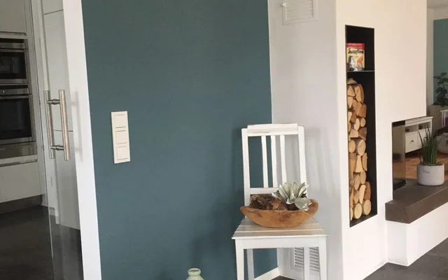 Moderner Wohnbereich mit dunkler Wand, weißem Stuhl, auf dem eine Holzschale mit Pflanzen steht, und einer Wandnische mit aufgestapeltem Kaminholz.