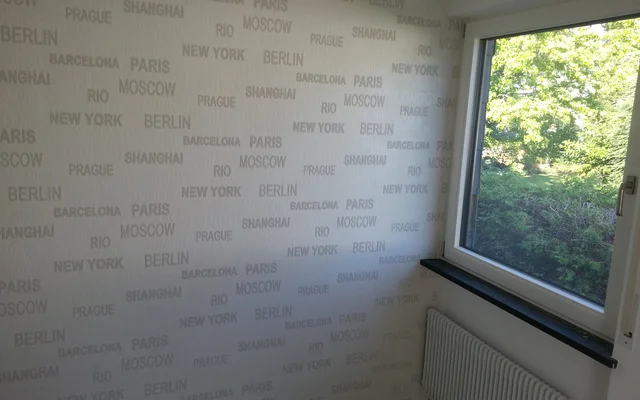 Fenster mit Blick auf grünes, sonniges Freiland und eine Wand mit Tapete, auf der verschiedene Städtenamen wie Berlin, New York, Paris und Shanghai wiederholt aufgedruckt sind.