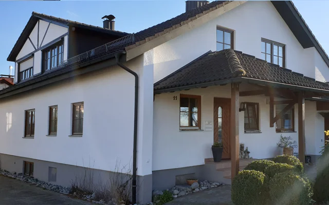 Ein weißes zweistöckiges Wohnhaus mit Holzveranda, Fenstern und einem kleinen Vorgarten mit Büschen bei Tageslicht.