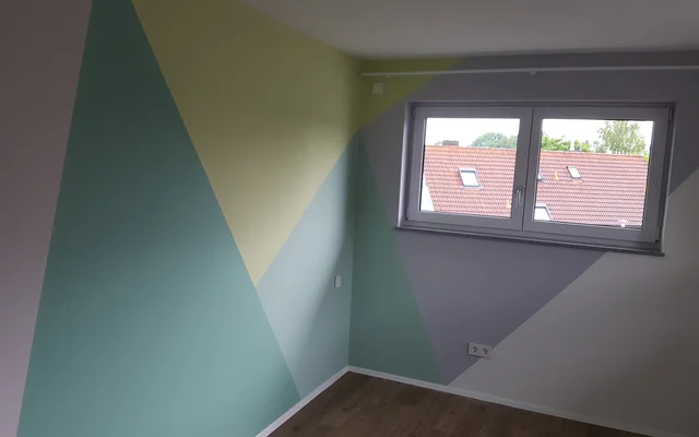 Zimmer mit leerem Holzboden, weiß gestrichenen und mit geometrischen Formen in Pastellfarben gestalteten Wänden, Fenster mit Blick auf Dachziegel von Nachbarhäusern.