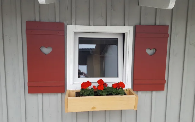 Kleines rechteckiges Fenster mit roten Klappläden, die Herzformen ausgeschnitten haben, darunter ein Holzkasten mit roten Blumen vor einer grauen Holzfassade.