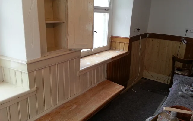 Raum mit hölzernen Sitzbänken entlang der Wände, einem kleinen offenen Schrank an der Wand und einem Fenster, das Tageslicht hereinlässt.