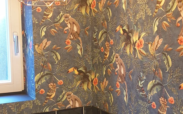 Badezimmer mit dunkelblauen floral gemusterten Tapeten und schwarzen, glänzenden Fliesen an der unteren Wand, beleuchtet von einer modernen, drahtigen Hängelampe mit sichtbarer Glühbirne neben einem Fenster.