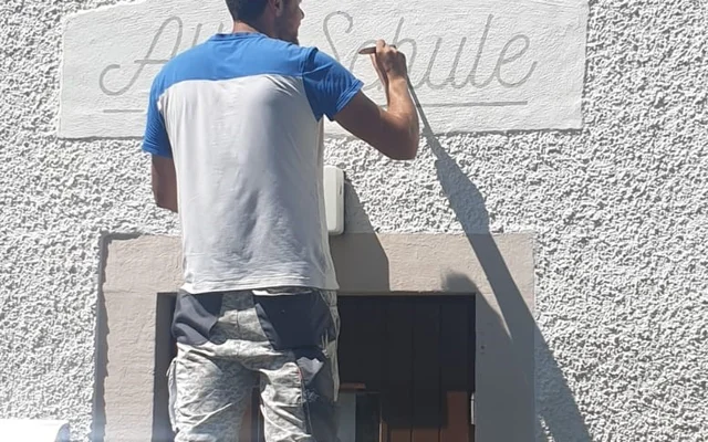 Ein Mann steht auf einer Leiter und zeichnet oder schreibt an einer weißen Wand über einer Tür.