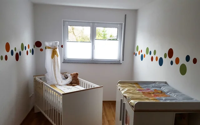 Kleines Babyzimmer mit Gitterbett und Teddybär, bunten Punkten an den Wänden und einer Wickelkommode unter dem Fenster.