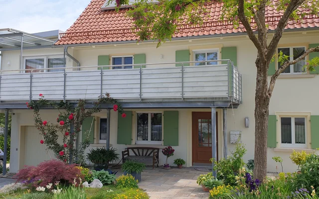 Modernes zweistöckiges Einfamilienhaus mit großem Balkon, gepflegtem Garten und blühenden Pflanzen im Vordergrund an einem sonnigen Tag.