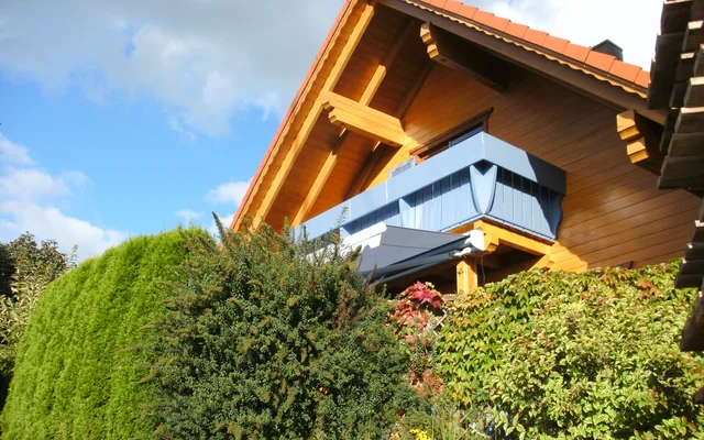 Haus mit Holzdach und Balkon hinter grünen Büschen und Hecken unter blauem Himmel mit Wolken.