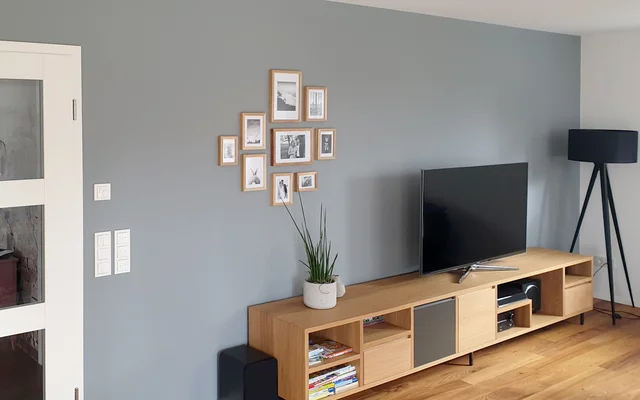 Moderner Wohnzimmerbereich mit grauer Wand, Holzsideboard, TV-Gerät, Stehlampe und mehreren gerahmten Fotos an der Wand.