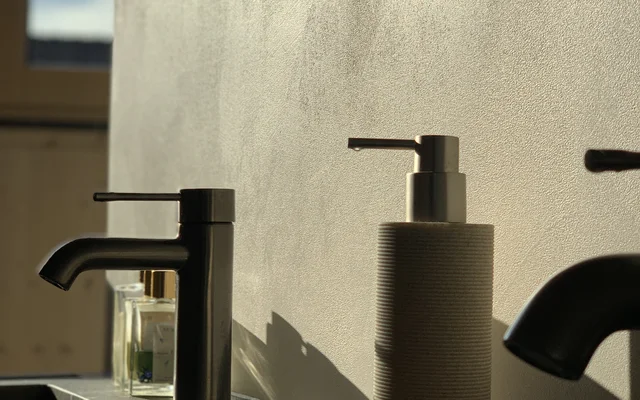 Nahaufnahme eines Waschbeckens mit zwei Wasserhähnen und einem Seifenspender vor einer hellen Wand mit Sonnenlicht, das Schatten wirft.