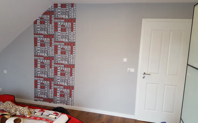 Schlafzimmer mit grauen Wänden, einer weißen Tür, einem Bett mit bunten Decken und einem Stück Wand mit Tapete, die Namen großer Städte wie Paris, New York und London zeigt.