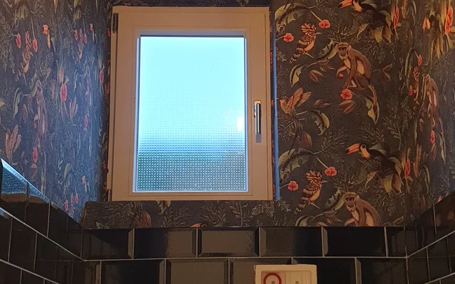 Badezimmer mit schwarzen Wandfliesen, dekorativer dunkler Tapete mit Blumenmuster, einem kleinen Fenster und einer modernen Hängelampe mit sichtbarer Glühbirne an der Decke.