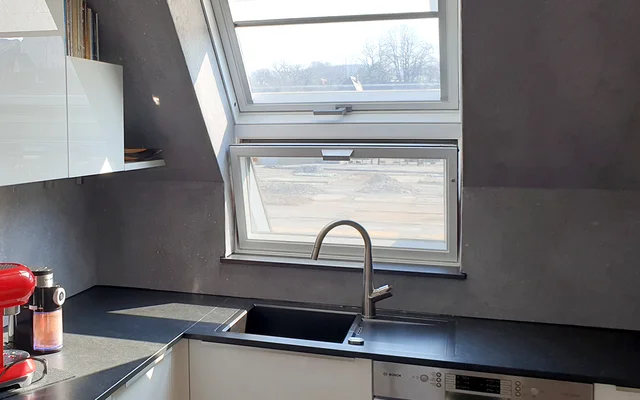 Helle Küche mit schwarzer Arbeitsplatte, Spüle unter einem geöffneten Dachfenster und verschiedenen Küchengeräten.