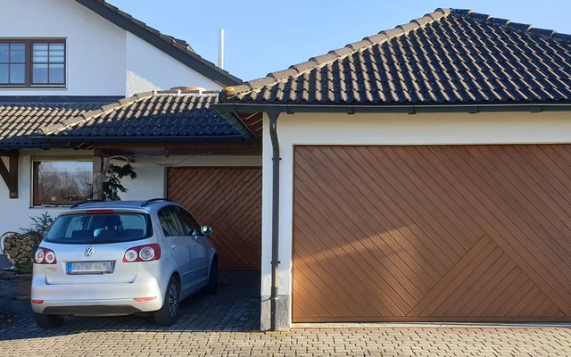 Ein silberner Volkswagen-Pkw steht in einer überdachten Einfahrt neben zwei großen, hölzernen Garagentoren an einem weißen Haus mit braunem Ziegeldach.