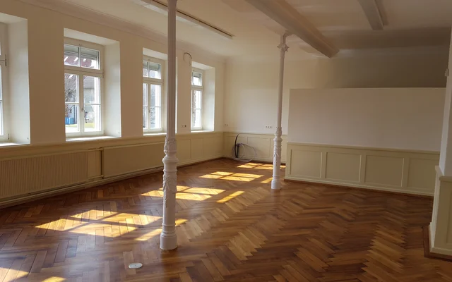 Leerer Raum mit Holzboden, vier großen Fenstern an einer Wand und zwei weißen Säulen in der Raummitte, Sonnenlicht fällt durch die Fenster auf den Boden.