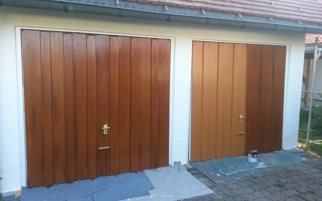 Zwei nebeneinander liegende Holztore mit goldfarbenen Türgriffen, links dunkler lackiert und rechts frisch mit hellbrauner Farbe gestrichen, vor einem weißen Gebäude mit rotem Ziegeldach. Auf dem Boden liegen Malerabdeckungen und ein Farbeimer.