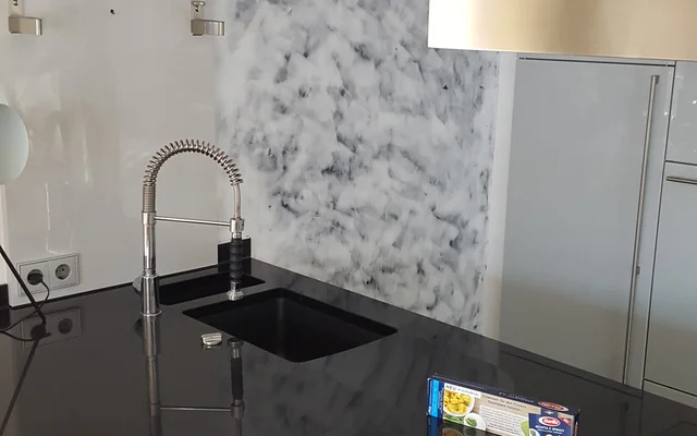 Moderne Küchenspüle mit schwarzer Arbeitsplatte, verchromtem Wasserhahn und Marmorwand im Hintergrund.