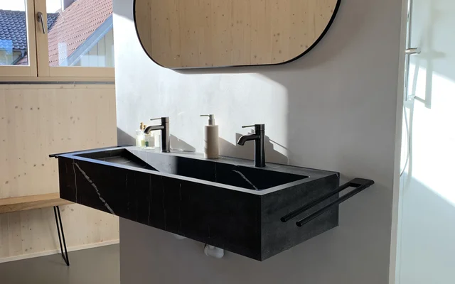 Modernes Badezimmer mit schwarzem Doppelwaschbecken, Wandspiegel mit abgerundeten Ecken und Fenster im Hintergrund, heller Holzverkleidung und minimalistischem Design.