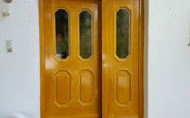 Holztür in gelblichem Farbton mit vier rechteckigen Fenstern, eingebaut in weiße Hauswand neben Fenster mit braunem Rahmen.