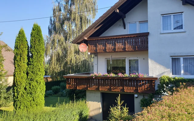 Zweistöckiges Wohnhaus mit weißen Wänden und braunem Holzbalkon, umgeben von grünen Bäumen und Sträuchern, unter klarem blauem Himmel.
