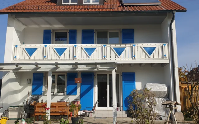 Mehrstöckiges Wohnhaus mit weißer Fassade, blauen Fensterläden und Balkon mit blau-weißen Geländern, Garten mit Möbeln und Pflanzen vor dem Haus.