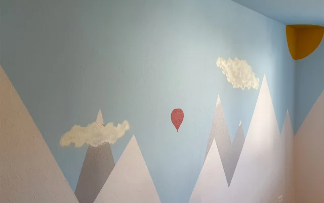 Wand mit gemalten Bergen, weißen Wolken, einem roten Heißluftballon und einer goldgelben Sonne in der Ecke.