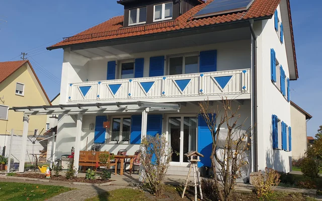 Mehrstöckiges weißes Haus mit blauen Fensterläden und Balkon, im Vordergrund Garten mit Terrasse, Möbeln und Gartengeräten, unter blauem Himmel an einem sonnigen Tag.