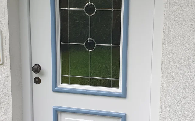 Weiße Haustür mit einem oberen, bogenförmigen Fenster mit Dekorglas und einem unteren, rechteckigen Paneel, beide mit blauer Umrahmung. Vor der Tür liegt eine schwarz-weiß gestreifte Fußmatte.