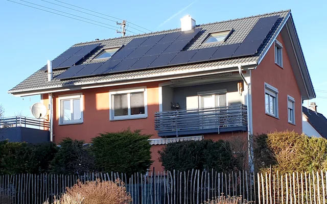 Zweistöckiges Wohnhaus mit oranger Fassade und Solarzellen auf dem Dach, umgeben von Hecken und einem Holzzaun.