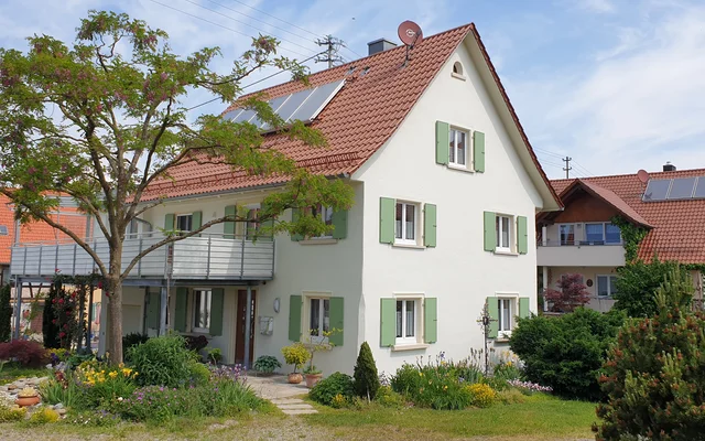 Modernes Einfamilienhaus mit weißer Fassade, grünen Fensterläden und Solarpanelen auf dem Dach, umgeben von Garten und Bäumen in einem Wohngebiet.