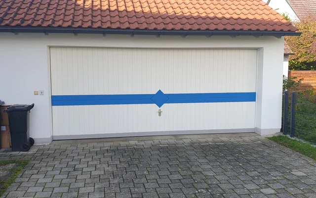 Geschlossene weiße Garagentür mit einem blauen Querstreifen und blauem Raute-Symbol, davor gepflasterte Einfahrt und zwei Mülltonnen links.