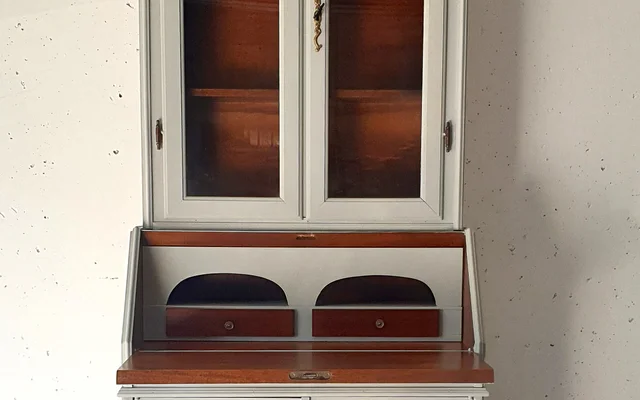 Heller Holzschrank mit Glastüren oben, offenem Schreibfach in der Mitte und geschlossenen Türen unten, steht vor einer weißen Wand neben einem kleinen Fenster.