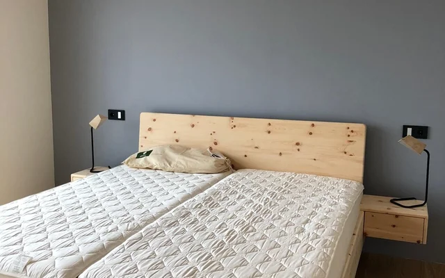 Schlafzimmer mit hellem Holzbett, zwei weißen Matratzen, einer grauen Wand im Hintergrund und je einer Nachttischlampe auf beiden Seiten des Betts.