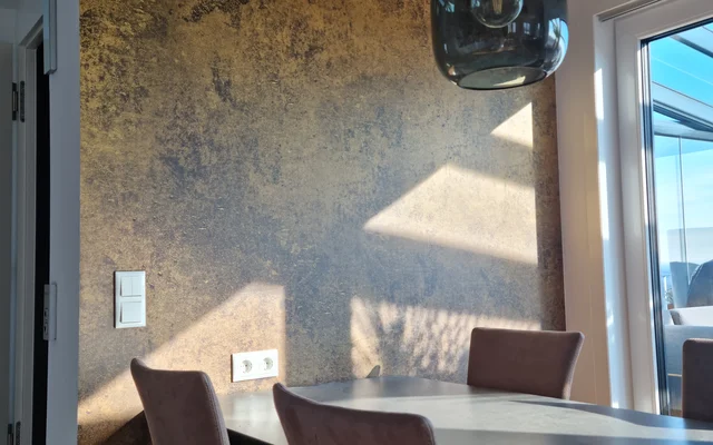 Essbereich mit vier braunen Stühlen um einen schwarzen Tisch, dahinter Wand mit rustikalem, braun-goldenem Muster, darüber moderne dunkelblaue Hängeleuchte, Licht fällt durch Fenster auf Wand und Tisch.