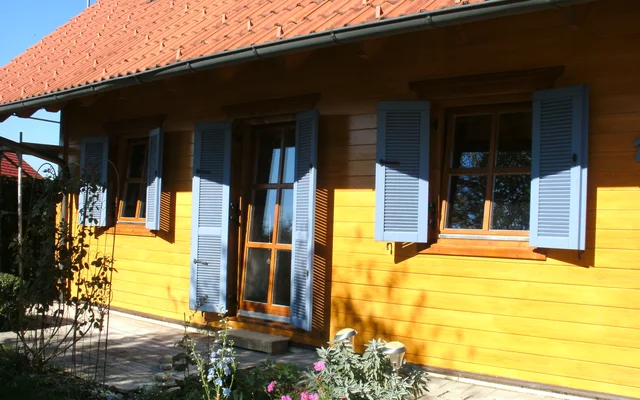 Holzhaus mit rot-gedecktem Dach, blauen Fensterläden und Tür auf einer sonnigen Terrasse mit kleinen Blumenbeeten davor.