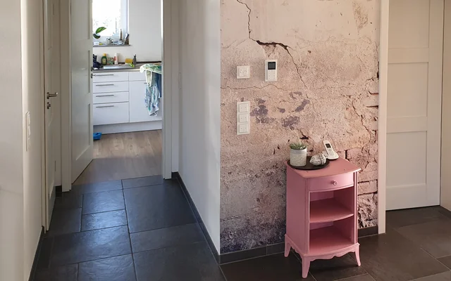 Flur mit dunklem Boden, kleiner rosa Kommode mit Pflanze und Telefon, sichtbar angrenzende Küche mit weißen Schränken und Fenster.