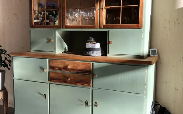 Grüner und brauner Küchenschrank mit Glasfenstern oben, gefüllt mit Gläsern und Flaschen, darunter geschlossene Schubladen und Türen, dekoriert mit einem kleinen Topfpflanze oben und einem Stoffbeutel daneben.