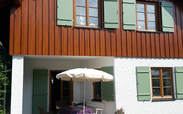 Haus mit Holzfassade im oberen Bereich und weißem Putz unten, Terrasse mit Tisch, Stühlen und Sonnenschirm, grüne Fensterläden, sonniger Tag.