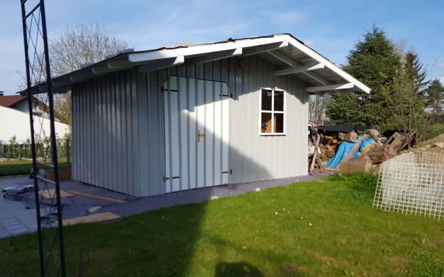 Kleines weißes Gartenhaus mit Holztür und Fenster auf einer Wiese im Sonnenschein, daneben Baumstämme und Bauaterialien.