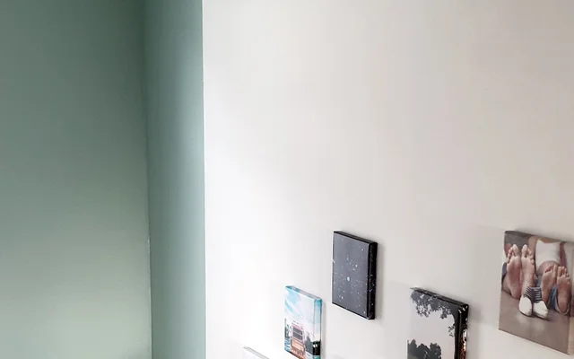 Weiße Wand mit mehreren kleinen quadratischen Bildern oder Fotoleinwänden, links angrenzend eine grün gestrichene Wand mit einem schmalen Fenster.