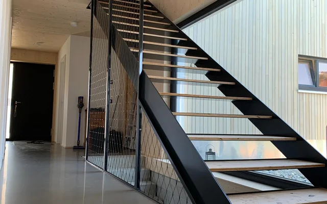 Moderner Innenraum mit schwarzer Metalltreppe, hellem Holz an Wand und Decke sowie großem Fenster, das Außenwand eines Nachbargebäudes zeigt.
