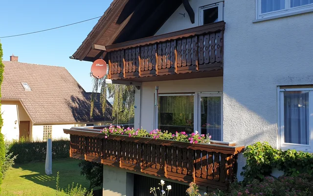 Haus mit Holzgeländerbalkon und Blumen, umgeben von grünem Garten und Bäumen bei sonnigem Wetter.