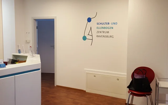 Empfangsbereich eines medizinischen Zentrums mit Stuhl, Tasche und Logo an der Wand.